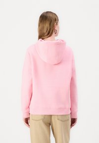 Donna con lunghi capelli castano chiaro, indossando un hoodie rosa e pantaloni beige, girata di spalle su uno sfondo bianco semplice.