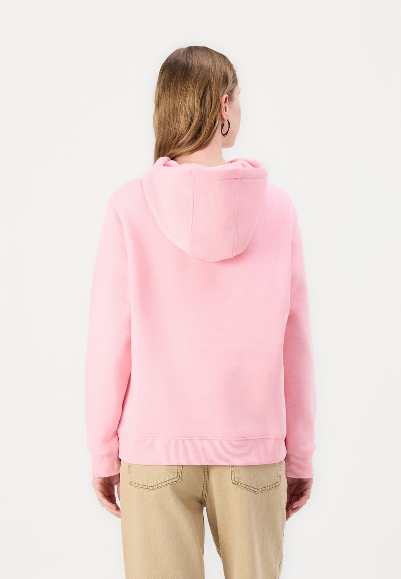 Donna con lunghi capelli castano chiaro, indossando un hoodie rosa e pantaloni beige, girata di spalle su uno sfondo bianco semplice.