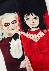 Los calcetines de punto presentan dos diseños: un personaje con cabello gris y rasgos oscuros sobre un fondo negro, y otro con una figura de cabello oscuro vestida de rojo.