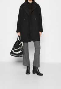Cappotto nero doppiopetto con cuciture bianche a contrasto, abbinato a pantaloni a quadri grigi e una borsa a tracolla a righe bianco e nera. Stivali neri alla caviglia.