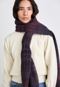 Wollschal in Dunkelblau und Lila mit einem karierten Muster. Weiche Textur mit fransigen Kanten. Über einem hellen Cremepullover getragen.