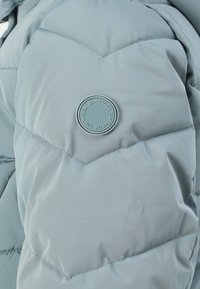 Lyseblå pufferjakke med chevron-syninger. Har en cirkulær logo-patch på ærmet. Materialet ser glat og quiltet ud.