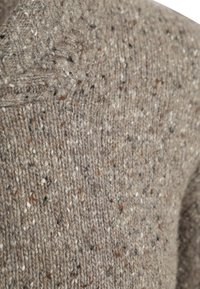 Bruine gebreide stof met een gespikkelde textuur, waarin vlekken van wit en donkere kleuren zijn verwerkt, wat een zachte, dikke uitstraling benadrukt.