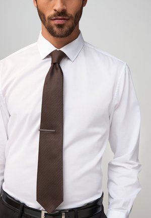 Man wearing a white dress shirt, brown textured necktie with a silver tie clip, and black belt against a light gray background.