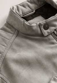 Col de veste en daim gris avec détails de panneaux cousus, bouton-pression métallique et fermeture éclair visible près du bord du col.