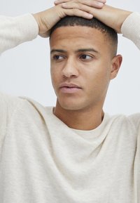 Suéter de punto color crema con un cuello redondo acanalado, tela texturizada y costuras visibles, usado por una persona con el pelo corto.