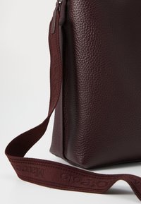 Sac en cuir bordeaux avec une surface texturée, doté d'une fermeture éclair et d'une bandoulière en tissu assortie avec des lettres en relief.