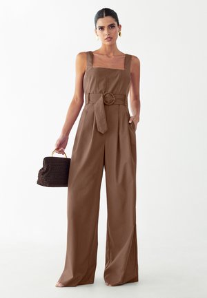 Kobieta ubraną w brązowy kombinezon z szerokimi nogawkami i paskiem, o kwadratowym dekolcie, trzymająca ciemną plecioną torebkę i nosząca złote kolczyki koła.