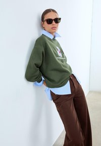 Sweatshirt vert olive avec un design de chien brodé, superposé sur une chemise à rayures bleues, associé à un pantalon large marron et des lunettes de soleil noires.