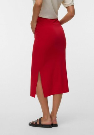 Kvinde iført en rød midinederdel med en slids bagpå, beige top og sorte slidesandaler, der står med den ene hånd på hoften.