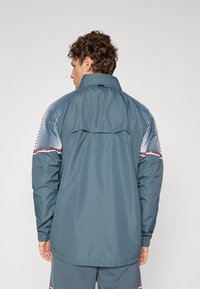 Herrenjacke in Dunkelgrau mit einem hohen Kragen, gemusterten Schulterpaneelen in Silber, roten Akzentstreifen und einem leicht verlängerten hinteren Saum.