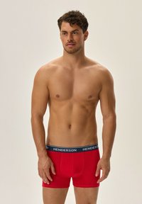 Czerwone bokserki z granatowym pasem, na którym widnieje napis "HENDERSON". Materiał wydaje się być miękki i elastyczny, dopasowany do sylwetki męskiego modela.