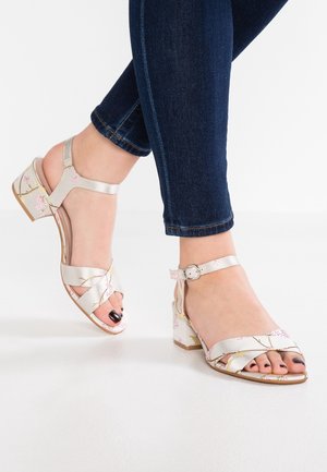 Fødder iført sølvfarvede blokhælede sandaler med blomsterremme rundt om anklerne, kombineret med mørkeblå skinny jeans på hvid baggrund.