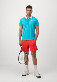 Sky blue polo shirt with white collar, red shorts, holding a tennis racket, white sneakers with colorful accents, and white socks.
