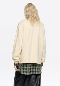 Krémová oversized mikina s dlhými rukávmi, so zošitým lemom, prehodená cez zelenú károvanú košeľu. Hladká látka s voľným strihom.