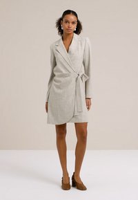 Donna che indossa un vestito avvolgente beige a maniche lunghe con cintura annodata, orecchini bianchi vistosi e scarpe marroni con tacco largo, in piedi su uno sfondo uniforme.