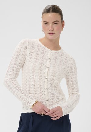 Vrouw met naar achteren gestyled haar, gouden oorringen, een wit gebreid vest met patroon en knopen, gouden armbanden en een marineblauwe broek.