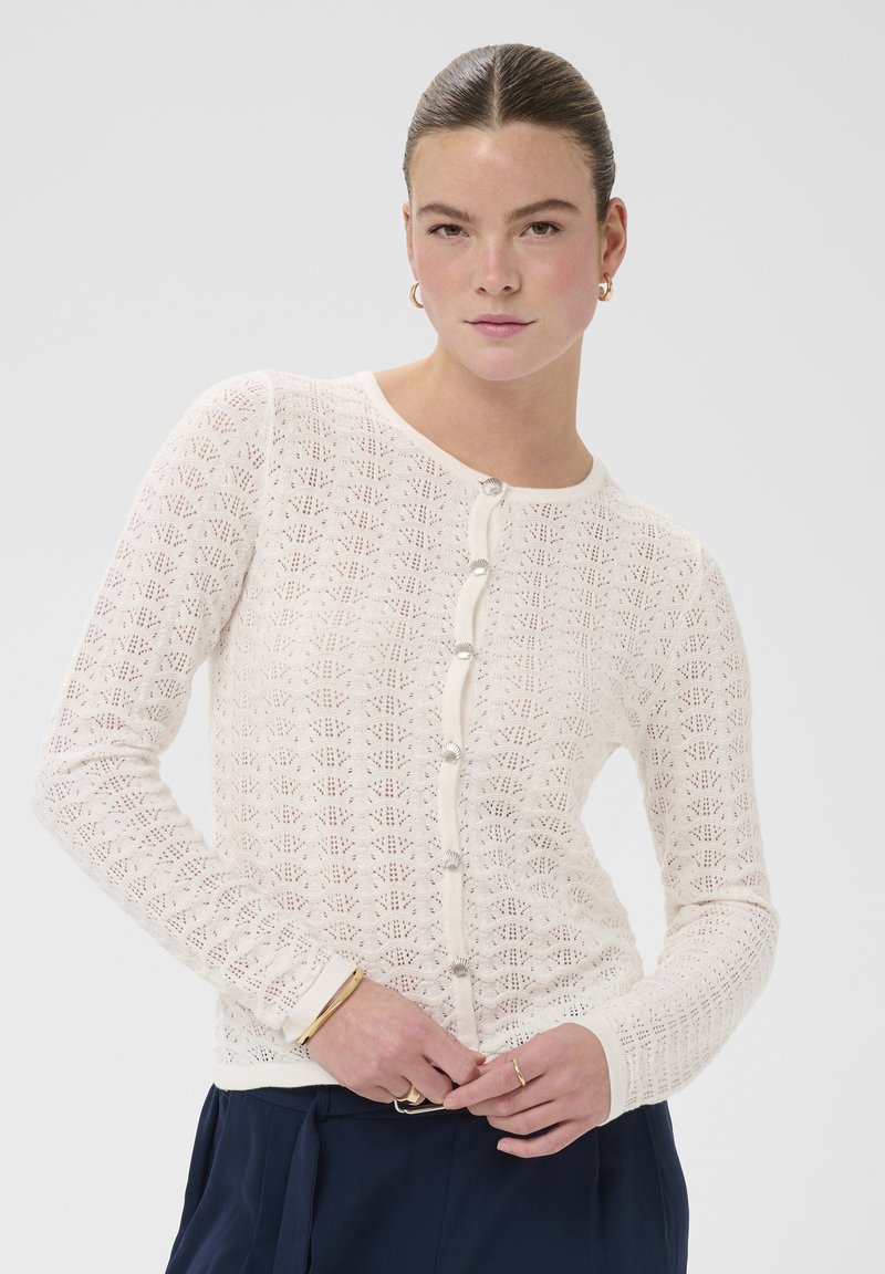 Femme aux cheveux tirés en arrière portant des boucles d'oreilles créoles dorées, un cardigan blanc à motifs boutonné, des bracelets dorés et un pantalon bleu marine.
