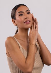 Goldarmband mit einem ovalen Charm, auf einem Handgelenk platziert. Modell mit langen Haaren, das ein beiges, geripptes Tanktop trägt, während die Hände sanft ihr Gesicht berühren.