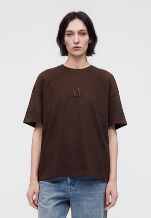 Jeune personne aux cheveux courts et foncés portant un t-shirt marron avec le logo A|X et un jean bleu, debout devant un fond clair uni.