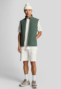 Grön ärmlös väst med dragkedja, texturerad vit skjorta, vita mönstrade shorts, beige sneakers och en krämfärgad mössa. Anmärkningsvärd gul logotyp på västen.