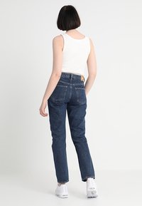 Jeans de mezclilla azul de talle alto con pierna recta, que presentan una sutil textura deslavada y bolsillos traseros tradicionales, combinados con una camiseta sin mangas blanca.
