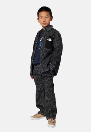 Ensemble de veste et pantalon à motifs noirs avec des accents blancs, affichant le logo "Kenzo Paris". Complété par des baskets beiges avec des détails graphiques.