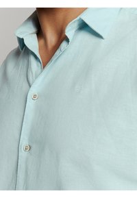 Close-up van een persoon die een lichtblauw button-up shirt draagt met een open kraag, waarbij de huid op de hals en bovenste borst zichtbaar is.