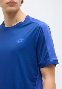 Νεαρό άτομο που φοράει μπλε αθλητική μπλούζα με κοντά μανίκια και μικρό λογότυπο στο στήθος σε απλό ανοιχτόφωτο φόντο.