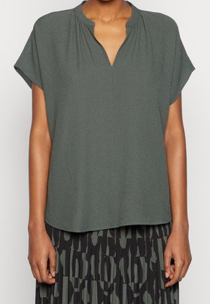 Vrouw draagt een losse, kortärmige, olijfgroene blouse met een V-hals, gecombineerd met een zwart-olijfgroen patroon rok.