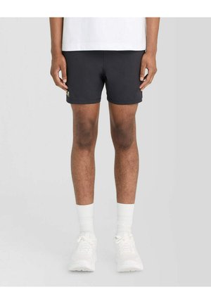 Mand iført sorte shorts, hvide sokker og hvide sneakers står op ad en ensartet lys grå baggrund.
