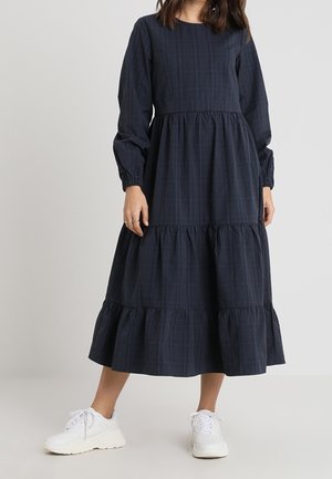 Kobieta w granatowej sukience midi z długim rękawem, z falbanami i delikatnym wzorem w kratę, w białych trampkach, stojąca na jednolitym tle.