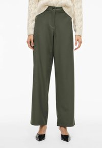 Olijfgroene broek met wijde pijpen, een gladde stof, een hoge taille met een knoop aan de voorkant en zijzakken, gecombineerd met zwarte puntige schoenen.