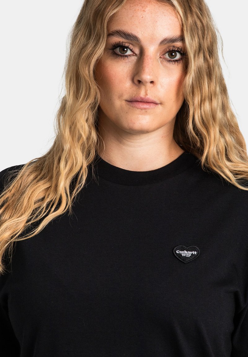 Schwarzes Langarmshirt mit rundem Halsausschnitt, featuring ein herzförmiges schwarz-weißes Logo im linken Brustbereich. Glatte Stofftextur.