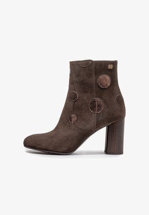 Botín de ante marrón con tacón bloque, decorado con parches circulares brillantes en el lateral y un pequeño logo dorado cerca de la parte superior.