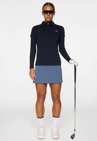 Kobieta ubrana w granatową golfową bluzkę z długim rękawem, niebieską spódnicę, okulary przeciwsłoneczne i białe rękawiczki, trzyma golfową pałkę.