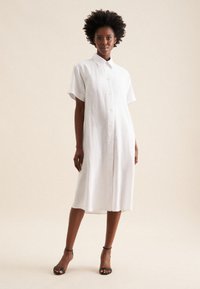 Weißes Leinenhemdkleid mit kurzen Ärmeln, vorderen Knöpfen, lässiger Passform, knielang, einfarbige Textur, kombiniert mit schwarzen Sandalen mit Riemen.