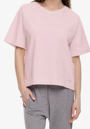 Kobieta w luźnym, jasnoróżowym T-shircie i szarych spodniach dresowych, z jedną ręką w kieszeni, na gładkim białym tle.