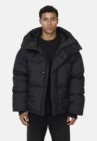 Giubbotto piumino nero caratterizzato da un design voluminoso, colletto alto e cappuccio staccabile. Tessuto liscio e lucido con chiusure a bottoni a pressione.