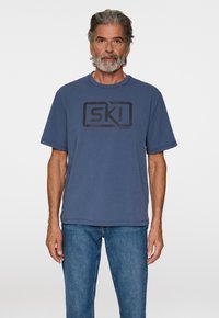 Sininen lyhythihainen t-paita pehmeästä puuvillasta, jossa on suuri musta "SKI" logo edessä. Yhdistetty farkkuhousujen kanssa.