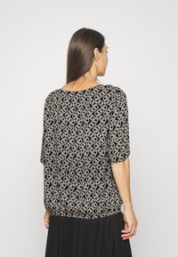 Top preto com um padrão geométrico de correntes em creme, com um corte solto, decote amplo e mangas de três quartos, feito de um tecido macio.