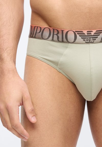 Άνδρας που φοράει ανοιχτό μπεζ εσώρουχο με καφέ ζώνη μέσης Emporio Armani, στέκεται με το αριστερό χέρι χαλαρό δίπλα στο μηρό.