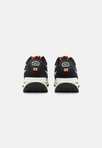Schwarze und cremefarbene Sneakers aus Wildleder und Mesh-Materialien. Mit orangefarbenen Akzentstreifen und einem Logo auf der Ferse. Dicke, strukturierte Sohlen.