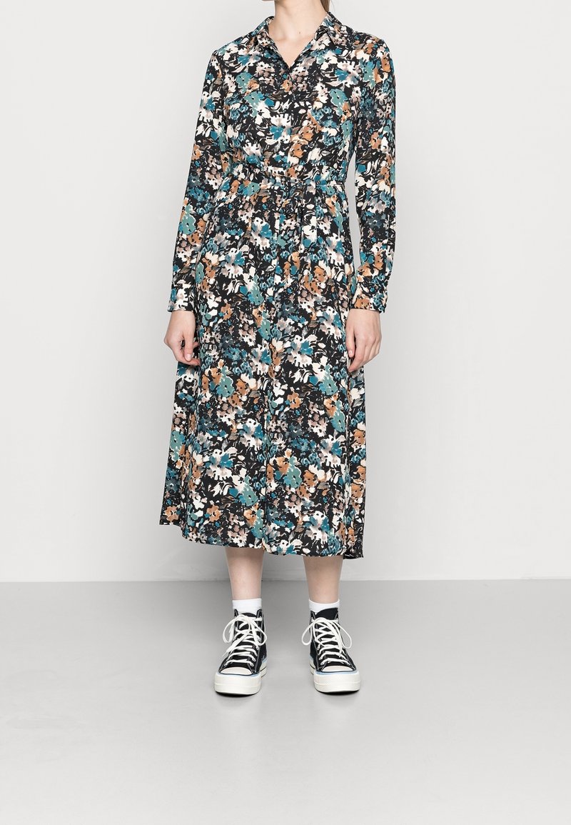 Robe à imprimé floral avec un fond noir, manches longues, col, taille cintrée et longueur midi. Associée à des baskets noires.