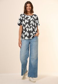 Sort-hvid mønstret top med korte ærmer, parret med brede lyse denim-jeans og cremede sneakers. Ensfarvet baggrund.