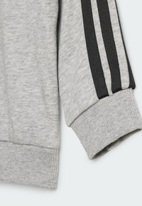 Grå sweatshirtsmaterial med ribbade muddar, med tre svarta ränder längs ärmen. Mjuk textur, avslappnad design och åtsittande fåll.