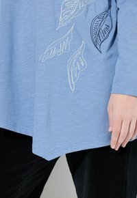 Ljusblå långärmad topp med bladbroderi i vitt och svart. Mjuk tyg, avslappnad passform, rundad nederkant, bärs med svarta kordbyxor.