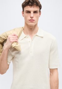 Jeune homme portant un polo crème texturé à manches courtes, tenant une veste beige sur son épaule droite, regardant droit devant.