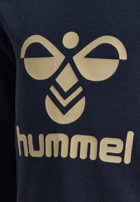 Un textile bleu marine présente un logo beige central avec des formes abstraites et le mot "hummel" dans une police grasse et arrondie en bas.