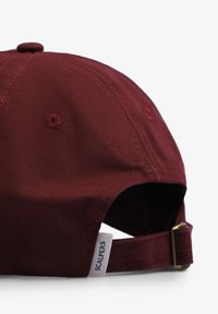 Gorra de algodón burdeos con un diseño bordado, visera curvada y correa ajustable con herrajes de metal, marcada con una etiqueta.